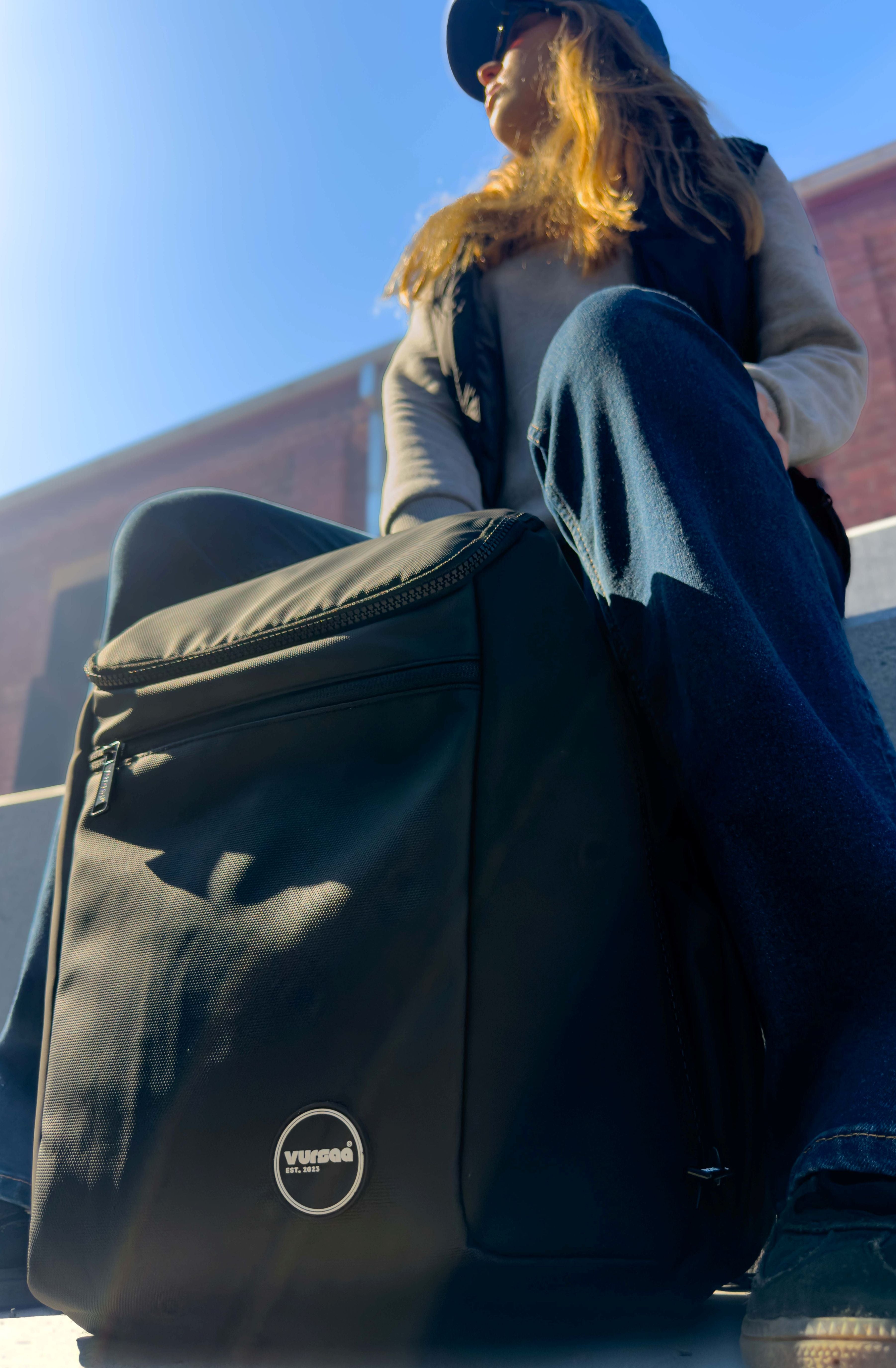 Person sitting on a ledge with a black modular backpack  featuring a logo, against a clear blue sky.
