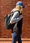 Person wearing a gray hoodie, black puffer vest, and blue cap against a brick wall. Carrying a modular black backpack