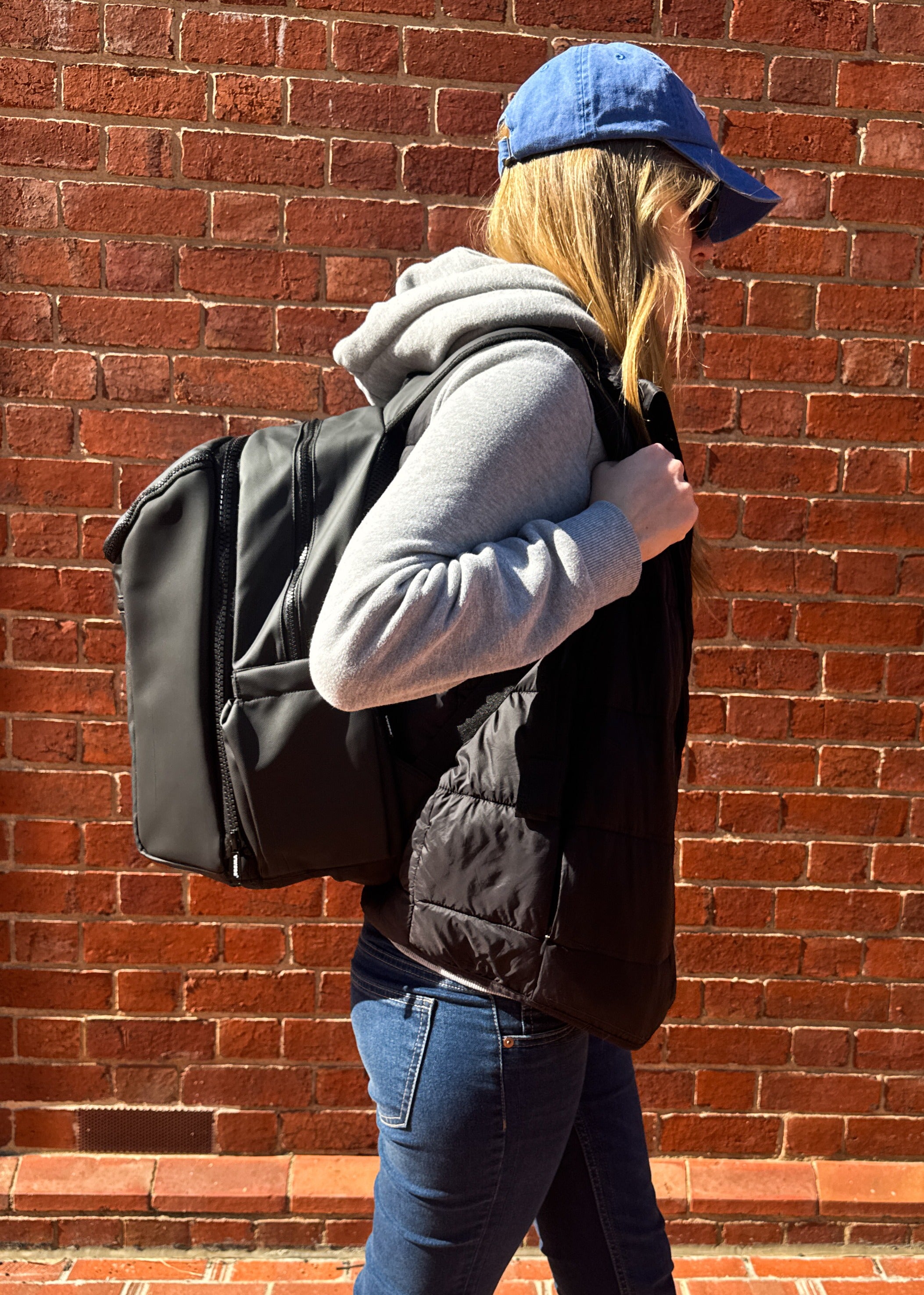Person wearing a gray hoodie, black puffer vest, and blue cap against a brick wall. Carrying a modular black backpack