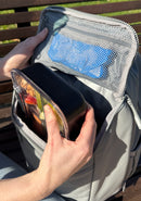 Person opening a gray insulated backpack with a black container and visible pocket holding an ice cooler. 