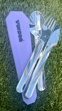 A set of stainless steel fork, knife and spoon with violet colored silicone case on grass.