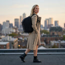 Woman in a trench coat with a vursaa backpack walking on a rooftop with a city skyline in the background