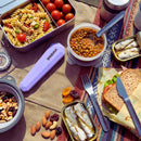 Assorted food items including pasta, beans, nuts, and a sandwich on a patterned tablecloth with travel utensils