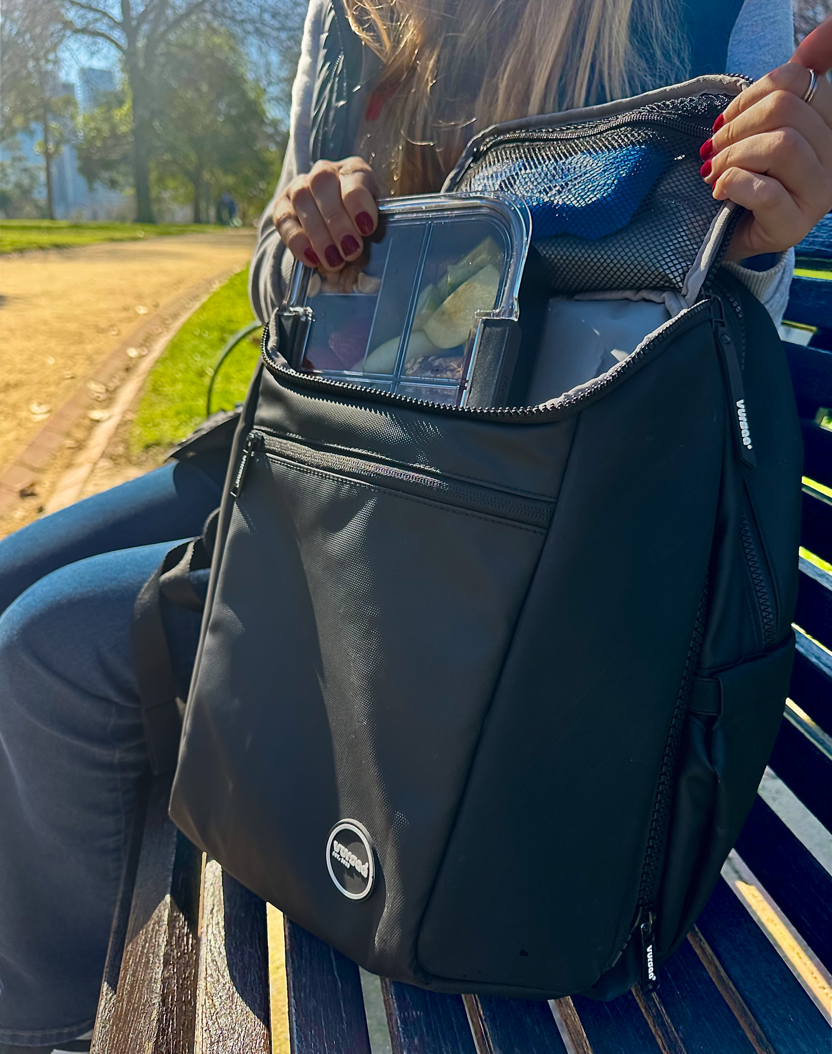 Person sitting on a bench with a black modular backpack exposing insulated front section, inside bag containing snacks. Logo in the corner