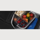 A clear plastic multi-compartment lunch box with a black lid, shown partially inside a black backpack. The compartments are filled with popcorn, tomatoes, blueberries, raspberries, and carrots, with a blue mesh pocket visible on the right.