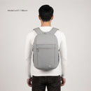 A man viewed from behind wearing a light gray backpack over a white long-sleeve shirt and dark pants, against a plain light gray studio background. Small text in the upper left reads 'Model is 6'1" / 185cm'.